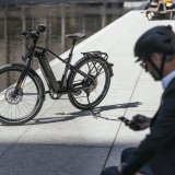 Ein E-Bike ist neben einer nassen Straße abgestellt. Im Vordergrund sitzt eine Person mit Fahrradhelm und schaut auf ihr Smartphone.