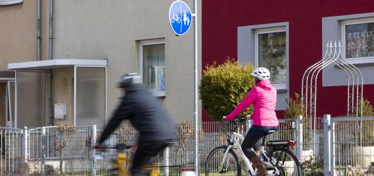 Zwei Personen fahren in dieselbe Richtung auf Fahrrädern durch die Stadt ? eine auf der Fahrbahn und eine auf dem geteilten Fuß- und Radweg.
