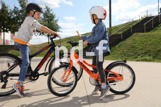 Zwei unterschiedlich große Kinder stehen mit Fahrrädern auf einem Platz und reden miteinander.