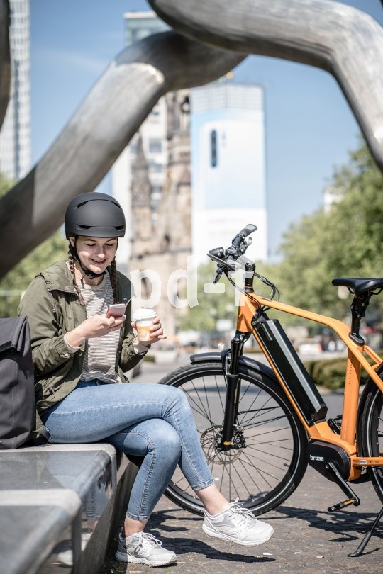 Eine Frau sitzt neben einem abgestellten E-Bike auf einer Bank und schaut lachend auf ein Smartphone.