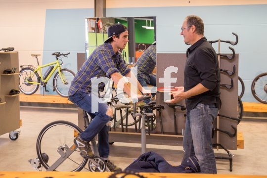 Ein Mann sitzt in einem Fahrradladen auf einem Ergometer. Ein zweiter Mann steht daneben und unterhält sich mit ihm.