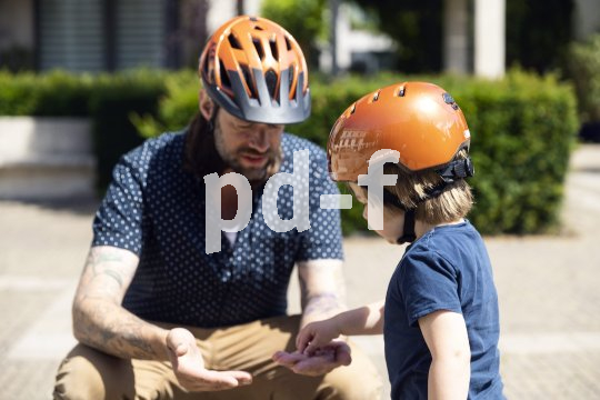 Ein Kind nimmt etwas aus der Hand eines Mannes, der vor ihm hockt. Beide tragen orange Fahrradhelme.