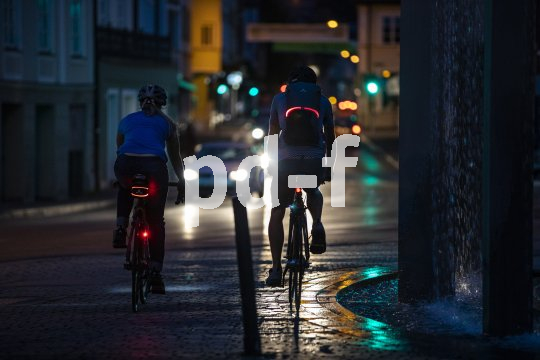 Zwei Personen fahren nachts auf Fahrrädern durch eine Innenstadt. An ihren Fahrrädern sind Rücklichter zu sehen und an ihren Rucksäcken und Taschen leuchtende oder reflektierende Elemente.