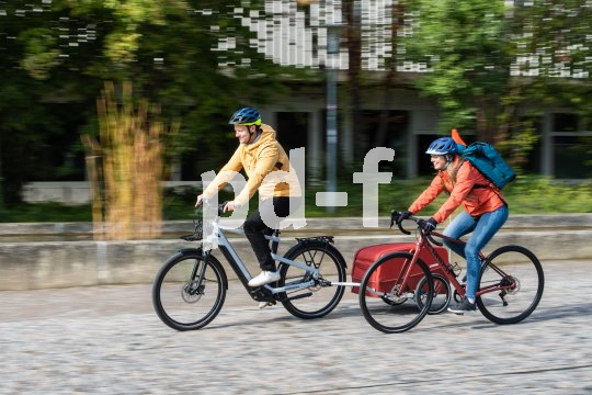 Ein Mann auf einem E-Bike mit Anhänger und eine Frau auf einem Rennrad fahren nebeneinander einen gepflasterten Weg entlang.