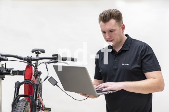 Ein Mann steht neben einem E-Bike in einem Montageständer. In der Hand hält er einen Laptop, der über Kabeln mit dem Rad verbunden ist.