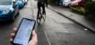 Eine Hand mit einem Smartphone, das einen Stadtplan anzeigt. Unscharf im Hintergrund ist eine Person auf einem Fahrrad in einem Wohngebiet zu sehen.