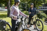 Zwei Frauen mit E-Bike stehen mit E-Bikes auf einem Radweg und lächeln einander zu.
