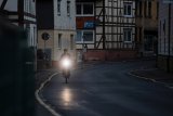 Ein Radfahrer fährt in der Dämmerung eine leere Straße zwischen Häusern entlang. Das Licht am Fahrrad leuchtet deutlich in die Kamera.