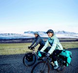 Eine Frau und ein Mann fahren lächelnd mit bepackten Fahrrädern vor einer entfernten Bergkulisse in offener Landschaft entlang.