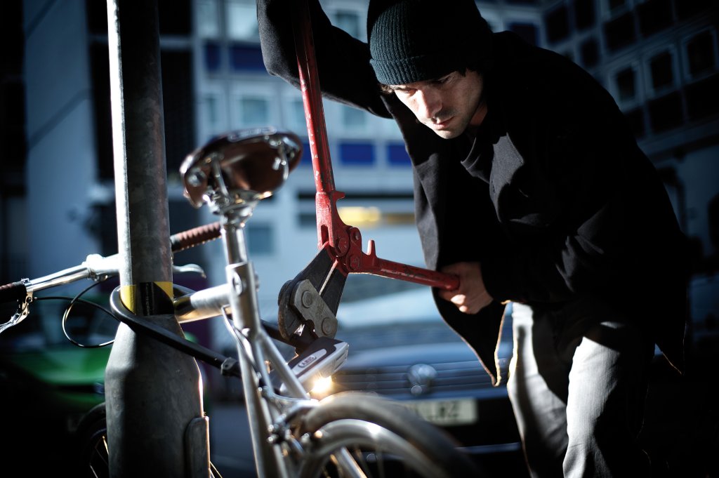 Ein Mann setzt einen Bolzenschneider an einem Bügelschloss an, mit dem ein Fahrrad an einem Metallpfosten gesichert ist.