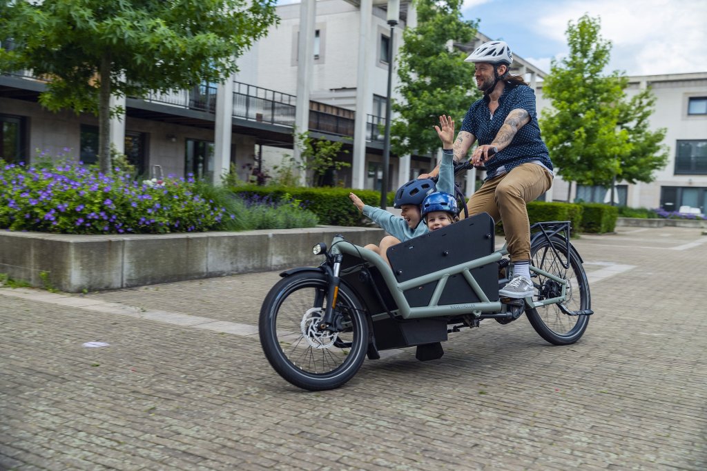 Ein Mann fährt auf einem Lastenrad mit zwei Kindern an Bord durch ein Wohngebiet.