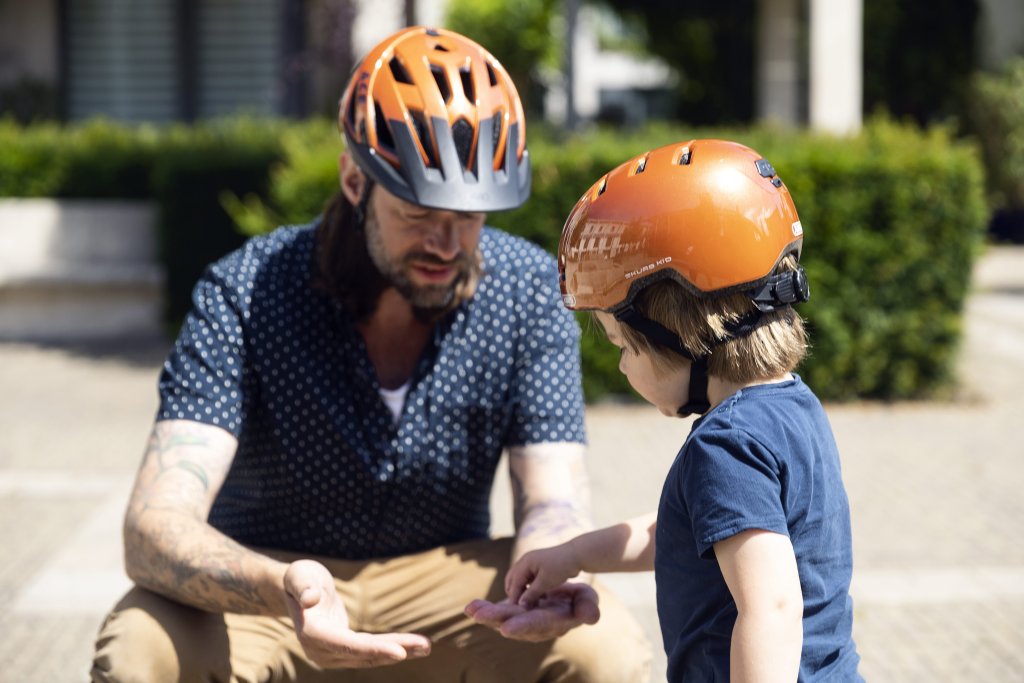 Ein Kind nimmt etwas aus der Hand eines Mannes, der vor ihm hockt. Beide tragen orange Fahrradhelme.