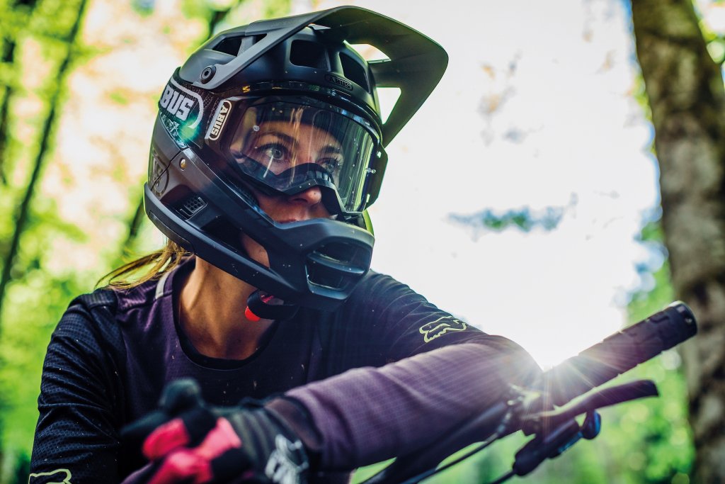 Frau mit Fullface-Helm mit Mountainbike im Wald bei Sonnenschein.