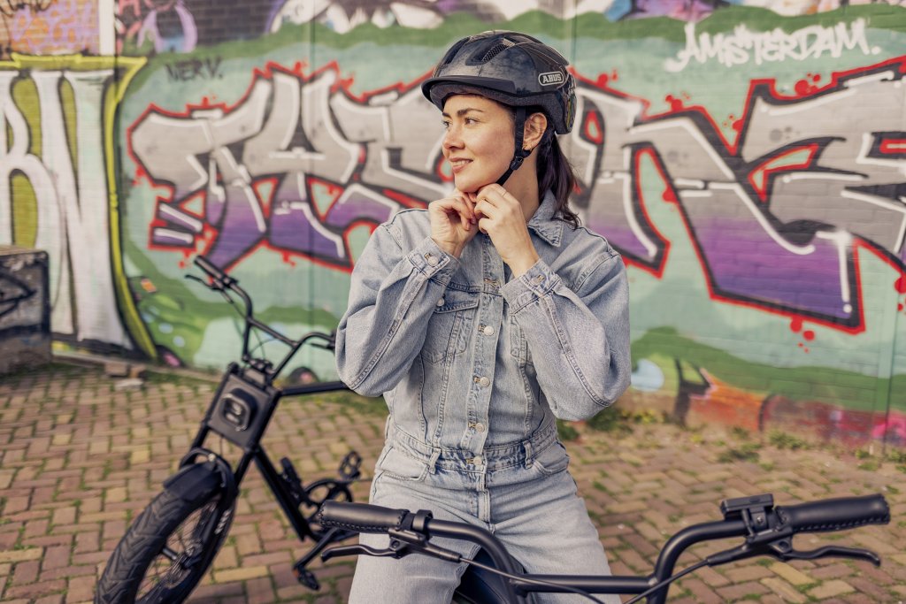 Eine Frau steht mit einem E-Bike vor einer Wand mit Graffiti und hantiert am Kinnriemen ihres Fahrradhelms.