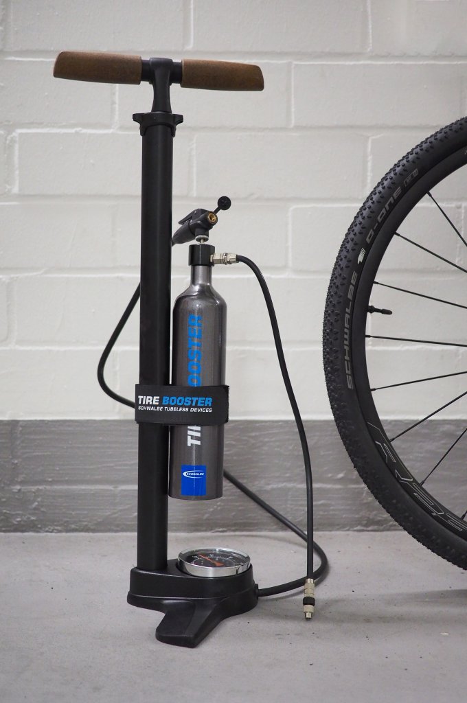 Neben einem Rad steht eine Fahrrad-Standpumpe mit einer Metallflasche und mehrere Schläuchen daran.