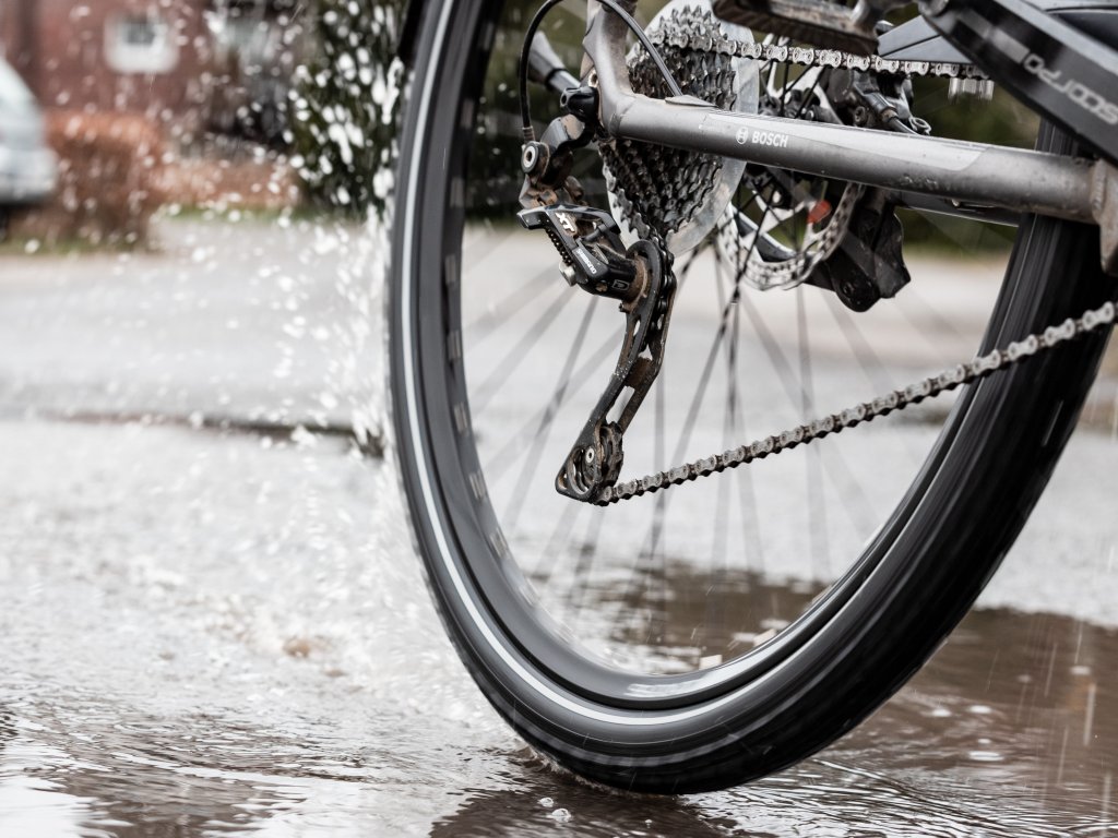 Bild des Hinterrades eines Fahrrades, dass durch eine Pfütze fährt. Vom Reifen spritzt Wasser.
