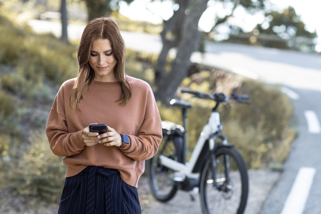 Eine Frau steht am Straßenrand vor einem Fahrrad und schaut auf ihr Smartphone.