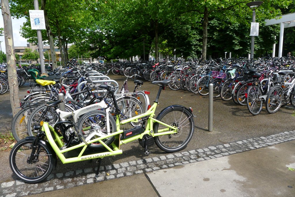 Ein Lastenrad mit einem Faltrad auf der Ladefläche, abgestellt am Rande einer großen Stellfläche für Fahrräder.