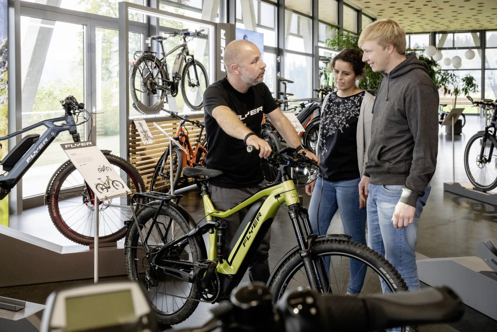 Ein Mann steht mit einem E-Bike in einem Fahrradladen und unterhält sich mit einer Frau und einem Mann.
