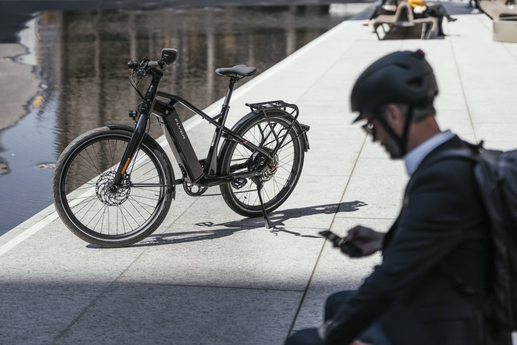 Ein E-Bike ist neben einer nassen Straße abgestellt. Im Vordergrund sitzt eine Person mit Fahrradhelm und schaut auf ihr Smartphone.