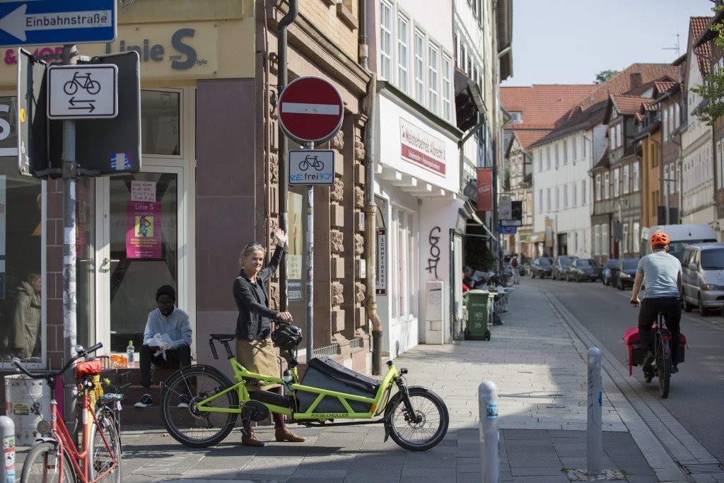 Eine Frau steht neben einem Lastenrad am Eingang einer innerstädtischen Einbahnstraße und zeigt auf das Verkehrsschild dazu. Ihr Fahrradhelm hängt am Lenker.