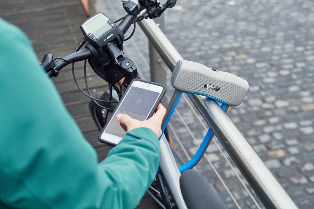 Blick über die Schulter einer Person, die ein Smartphone vor eine Bügelschloss hält, mit dem ein E-Bike an ein Geländer angeschlossen ist.