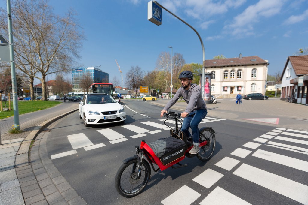 Ein Lastenradfahrer überquert eine Straße innerhalb einer Fahrbahnmarkierung. Ein Auto wartet im Hintergrund.