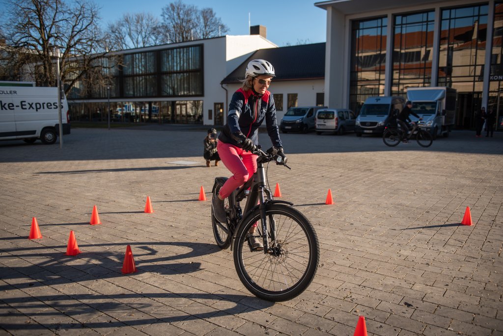 Eine Person fährt auf einem Platz mit einem E-Bike zwischen orangefarbenen Pylonen.
