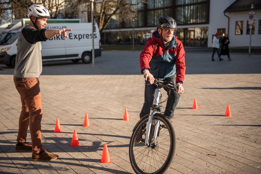 Ein Mann fährt mit einem E-Bike zwischen orangefarbenen Hütchen während ein zweiter Mann daneben steht und ihm die Richtung weist.