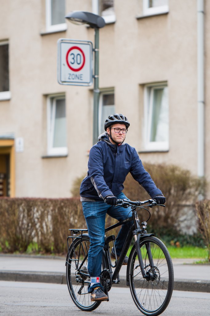 Ein Mann fährt auf einem Fahrrad auf einer Straße. Im Hintergrund ist vor einem Wohnhaus ein Verkehrsschild mit Geschwindigkeitsbegrenzung 30 km/h zu sehen.