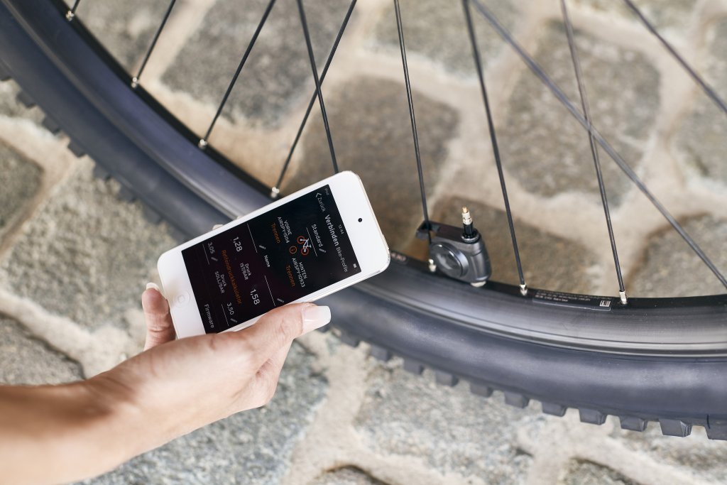 Eine Person hält ein Smartphone vor einen Fahrradreifen, an dessen Ventil ein kleines, schwarzes Gerät montiert ist.