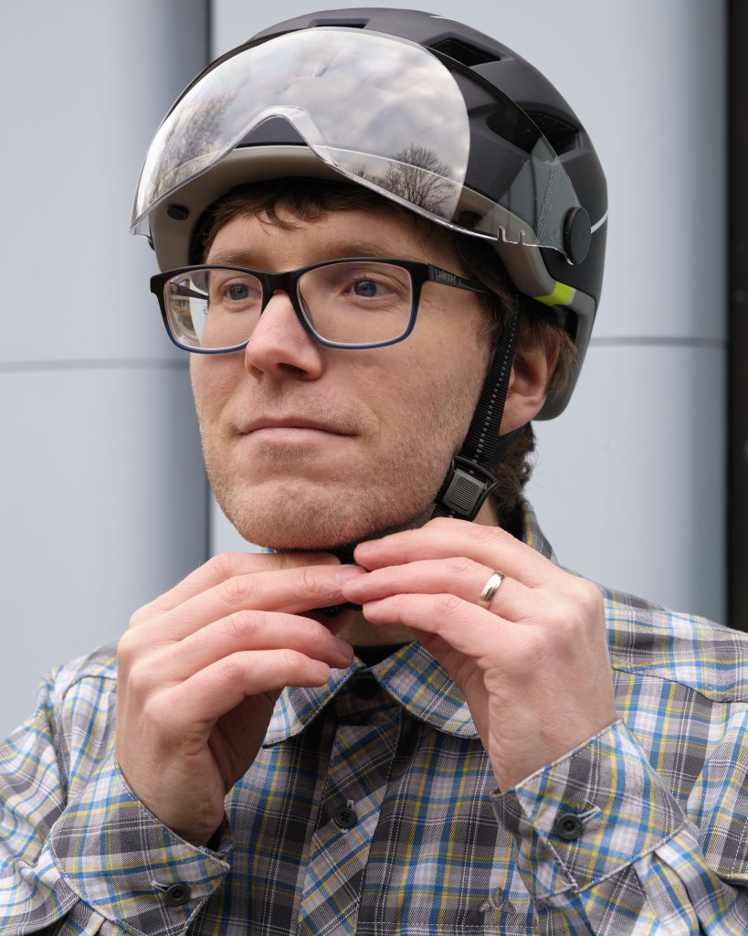 Ein Mann mit Fahrradhelm bedient den Verschluss des Helmriemens unterm Kinn mit beiden Händen.