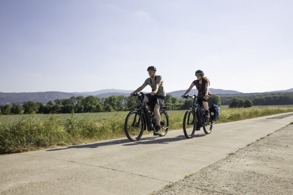 Zwei Personen fahren auf E-Bikes einen betonierten Weg neben einem Feld entlang.