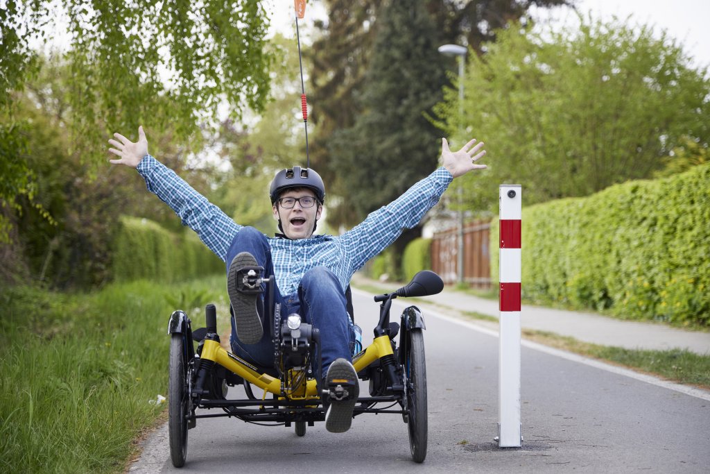 Ein Mann sitzt in einem dreirädrigen Liegerad neben einem Poller auf einem Radweg und streckt freudig die Hände nach oben.