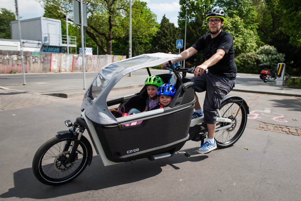 Ein Mann fährt ein Lastenrad mit zwei Kindern und Regenverdeck.