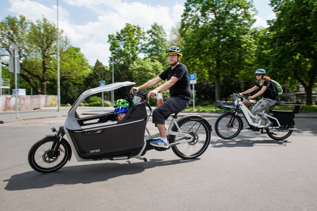 Ein Mann fährt ein Lastenrad mit zwei Kindern an Bord. Hinter ihm ist eine Frau auf einem E-Bike unterwegs.