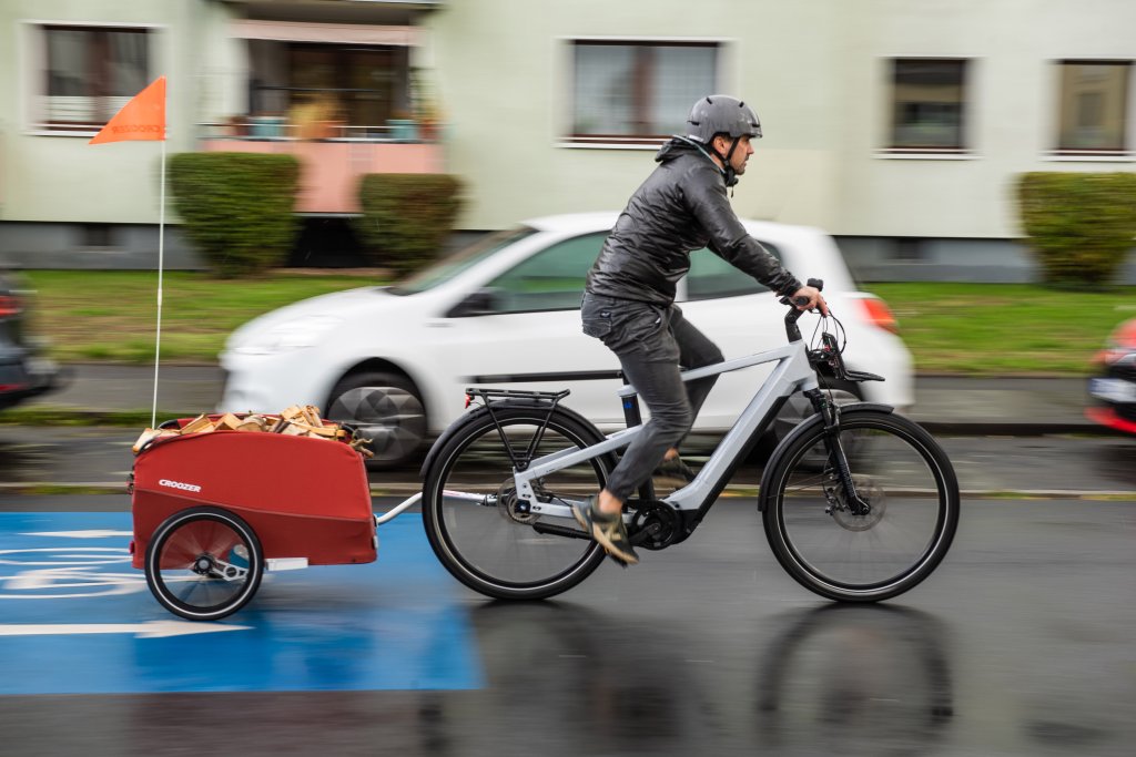 Ein Mann fährt auf einer Fahrradstraße mit einem E-Bike durch ein Wohngebiet. Am Rad hängt ein roter Anhänger.