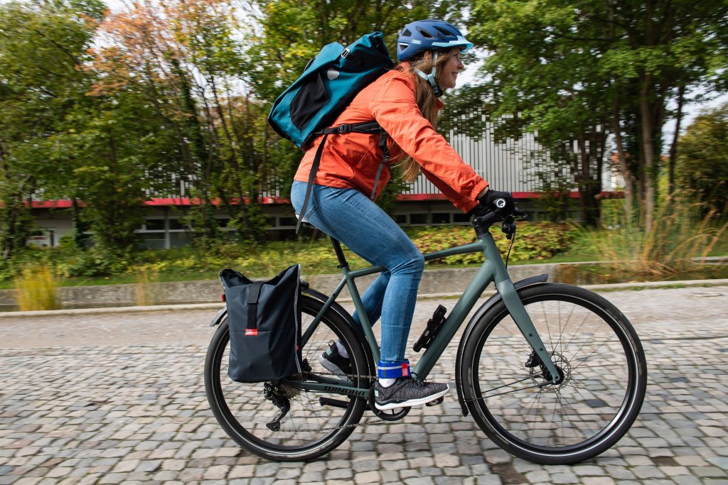 Eine Radfahrerin mit Rucksack und Gepäcktasche am Fahrrad fährt eine Straße mit Kopfsteinpflaster entlang.