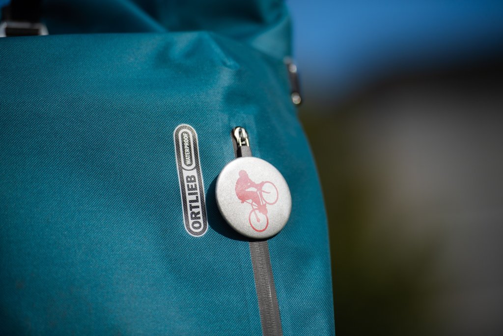 Ein runder Button mit eine:r stilisierten Radfahrer:in an einer Tasche mit dem Logo der Firma Ortlieb.