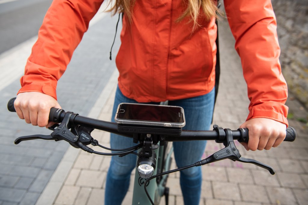 Ein Smartphone ist mittig auf dem Lenker eines Fahrrades montiert. Eine Person mit roter Jacke greift den Lenker.