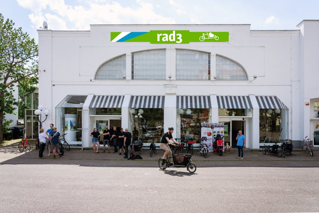 Mehrere Personen stehen vor einem Ladengeschäft mit dem Schriftzug 'rad3'. Ein Mann fährt mit einem Lastenrad los.
