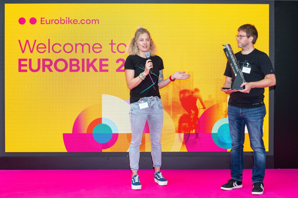 Eine Frau und ein Mann stehen auf einer Bühne auf er Eurobike und zeigen einen Fahrradakku.