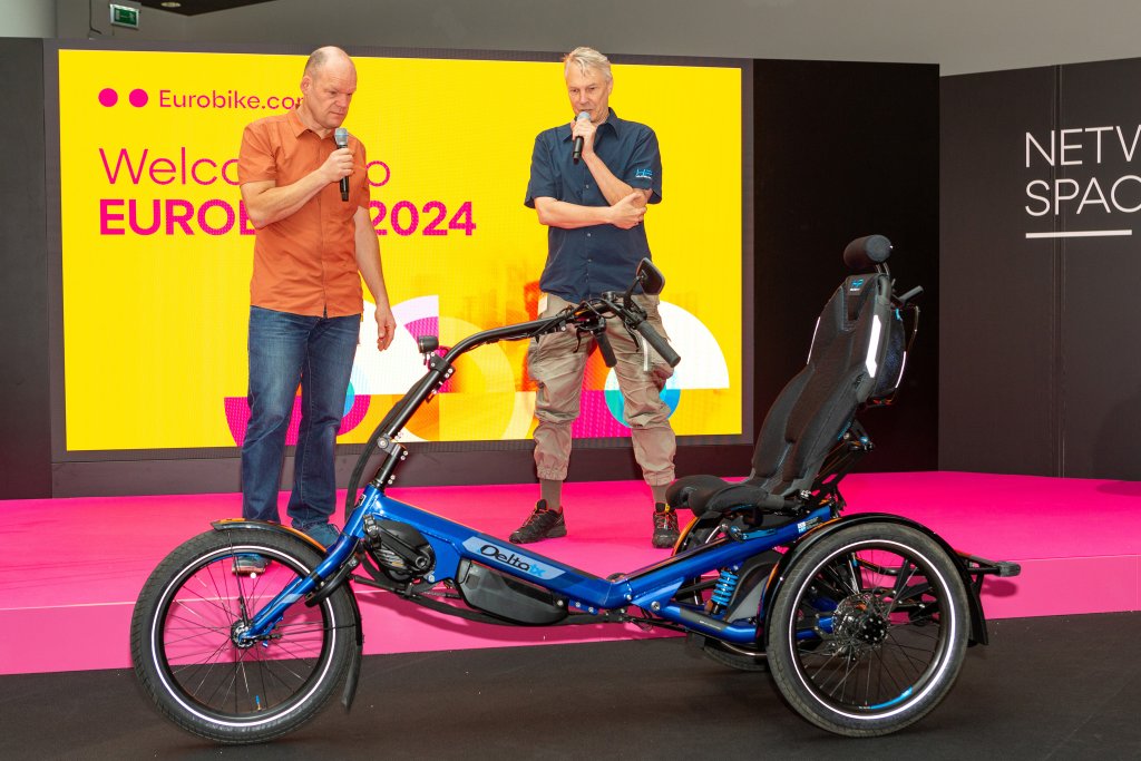 Zwei Männer auf einer Bühne auf der Eurobike stehen vor einem Sesseldreirad.