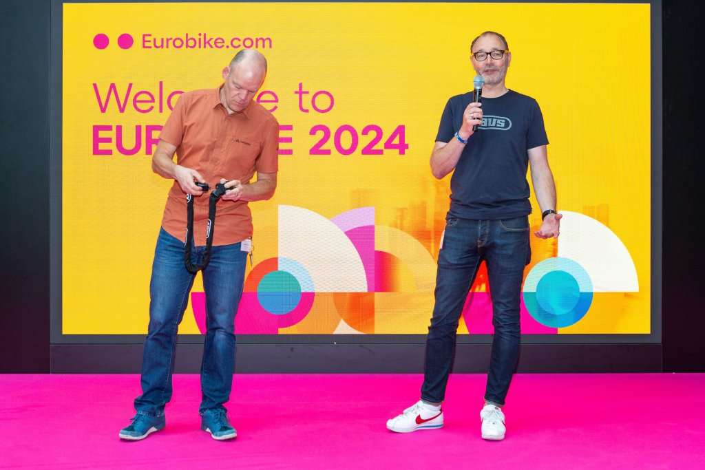 Zwei Männer zeigen auf einer Bühne auf der Eurobike ein Fahrradschloss.