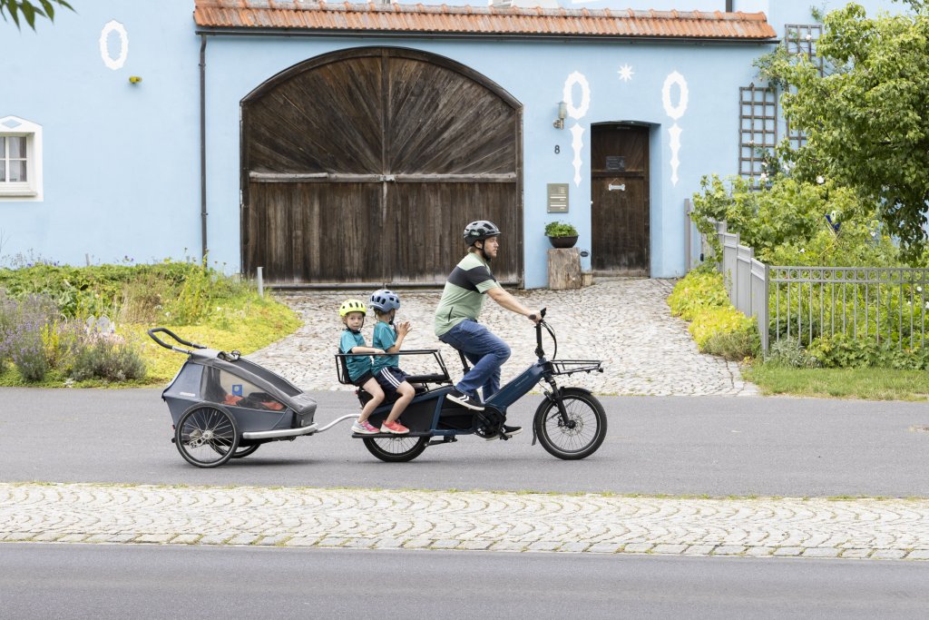 Mann fährt mit Rad und zwei Kindern plus Anhänger.