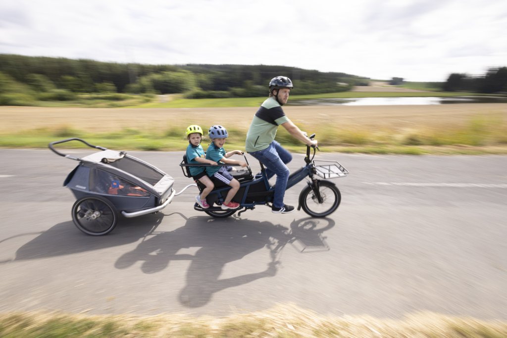 Mann fährt mit Longtail-Lastenrad und zwei Kindern sowie Anhänger