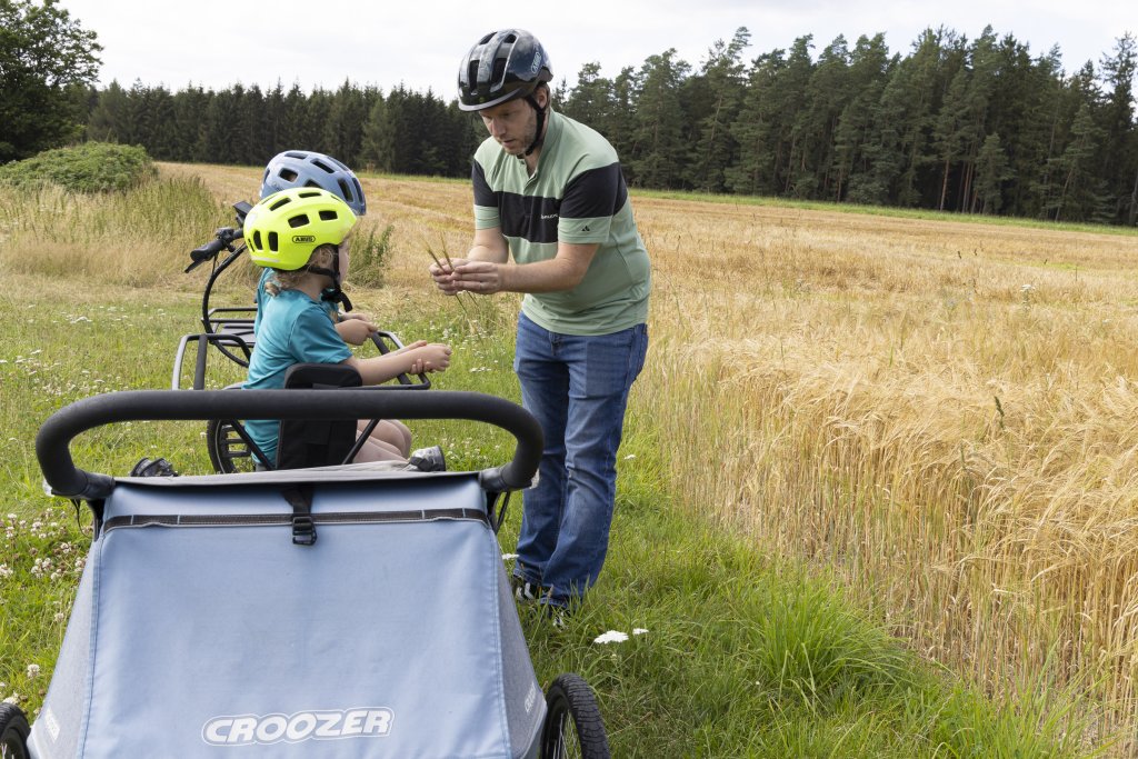 Mann zeigt Getreide an zwei Kinder, die auf Lastenrad sitzen.