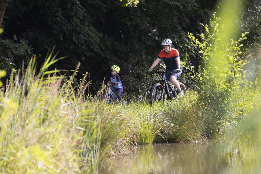 Mann und Kind fahren mit Fahrrad an Wasser entlang.