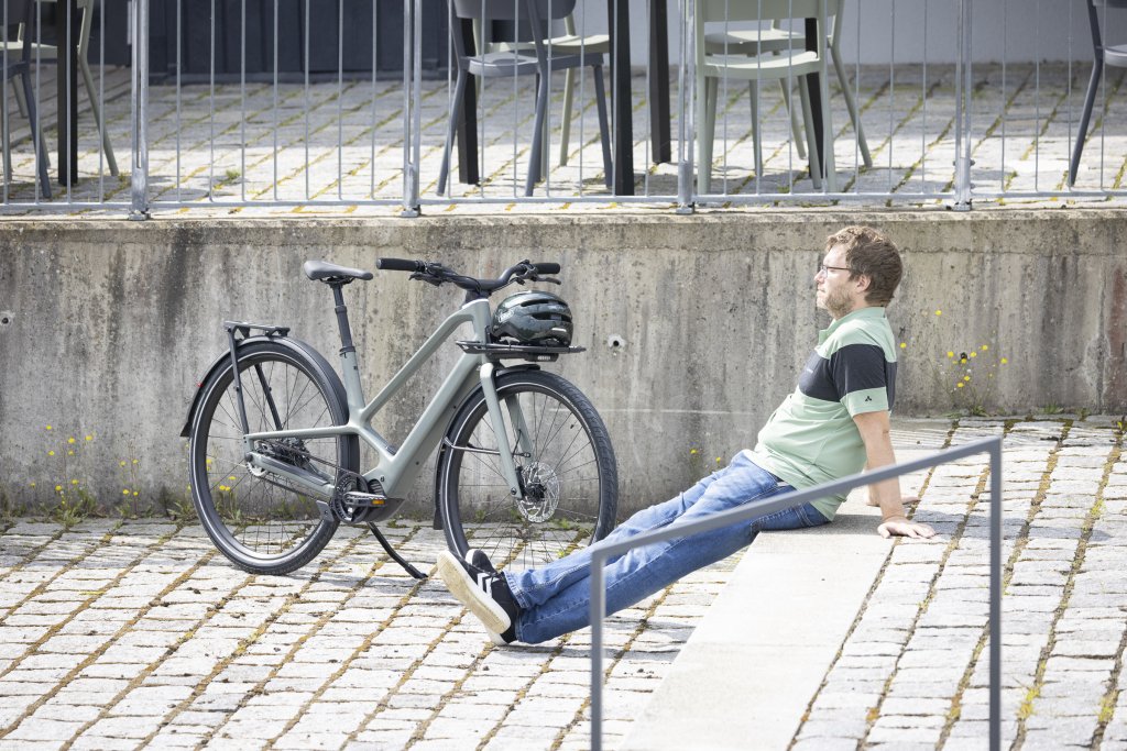 Mann sitzt auf Stufe und neben ihn steht ein E-Bike vor Betonmauer.