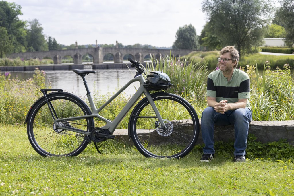 Mann sitzt auf Stein und hat neben sich eine E-Bike stehen. Im Hintergrund sieht man eine alte Steinbrücke.